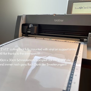 May include: A gray Brother ScanNCut DX cutting machine with a 12" x 12" cutting mat fully mounted with vinyl on support arms. The text reads "12" x 12" cutting mat fully mounted with vinyl on support arms still flat thanks to the extension!!! 30cm x 30cm Schneidematte komplett voll mit Vinyl-Folie und immer noch ganz flach Dank der Erweiterungen!!"