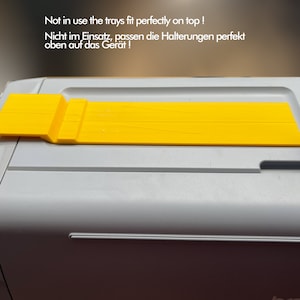 May include: A yellow plastic tray with a raised edge on one side. The tray is sitting on top of a gray machine. The text "Not in use the trays fit perfectly on top! Nicht im Einsatz, passen die Halterungen perfekt oben auf das Ger&auml;t!" is visible in the image.