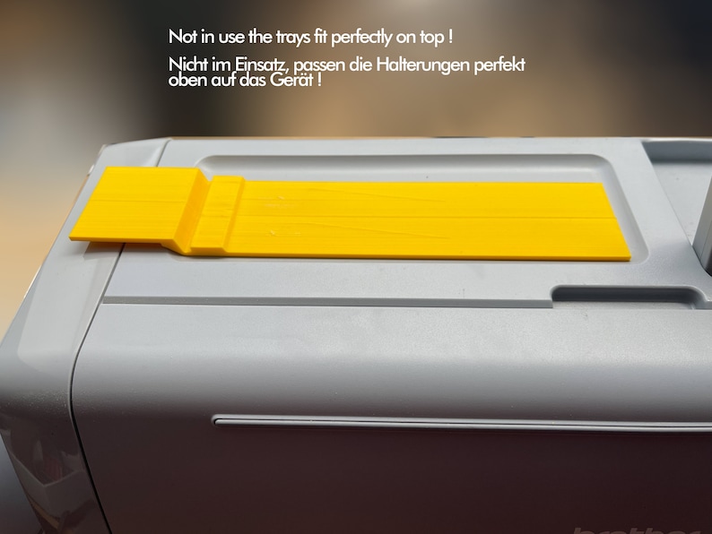 May include: A yellow plastic tray with a raised edge on one side. The tray is sitting on top of a gray machine. The text "Not in use the trays fit perfectly on top! Nicht im Einsatz, passen die Halterungen perfekt oben auf das Ger&auml;t!" is visible in the image.