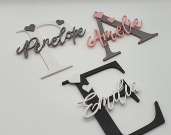 May include: Three wooden letters, P, A, and E, painted in white, gray, and black, with the names Penelope, Amelie, and Emilia written in white on each letter. Each letter has small pink or gray hearts attached.