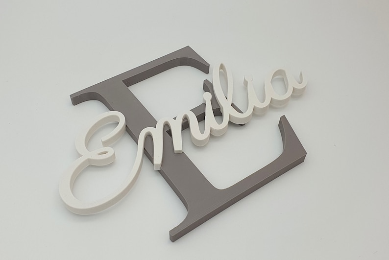 May include: A white wooden name sign with the name "Emilia" in cursive script. The name is attached to a larger gray wooden letter "E".