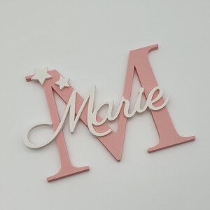May include: Pink wooden letter M with the name "Marie" written in white on it. Two white stars are on the top of the letter.