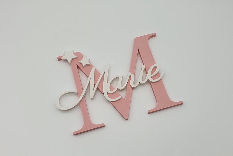 May include: Pink wooden letter M with the name "Marie" written in white on it. Two white stars are on the top of the letter.