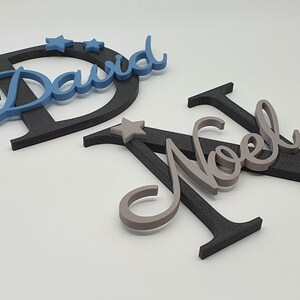 May include: Two names, "David" and "Noel", are spelled out in cursive script with black and gray letters. The letters are layered on top of each other and have a 3D effect. There are small gray stars scattered around the letters.