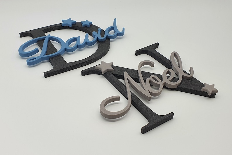 May include: Two names, "David" and "Noel", are spelled out in cursive script with black and gray letters. The letters are layered on top of each other and have a 3D effect. There are small gray stars scattered around the letters.