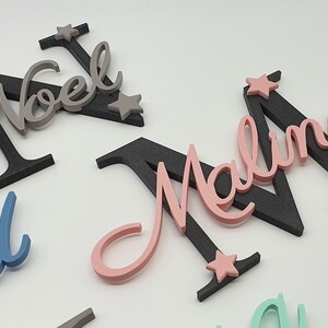 May include: Three wooden names with black backing and star accents. The names are Noel, Malina, and David. The names are in different colors: gray, pink, and blue.