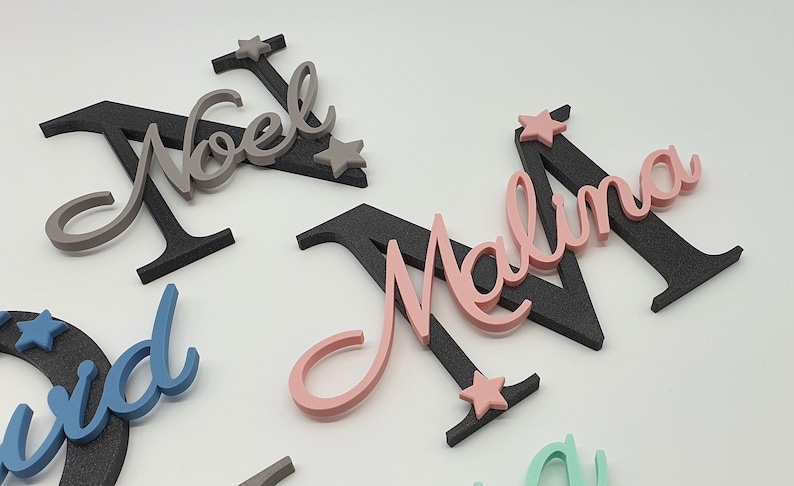 May include: Three wooden names with black backing and star accents. The names are Noel, Malina, and David. The names are in different colors: gray, pink, and blue.