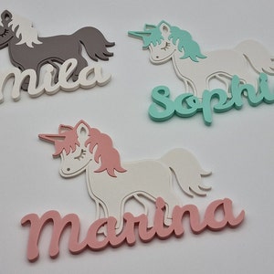 May include: Three wooden unicorn name plaques with the names Mila, Sophie, and Marina. The unicorns are white with pink, teal, and gray manes and tails. The names are in white, teal, and pink.
