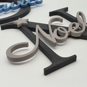 May include: Gray wooden letters spelling "Noel" with two small gray stars. The letters are layered on top of a black wooden letter "A".