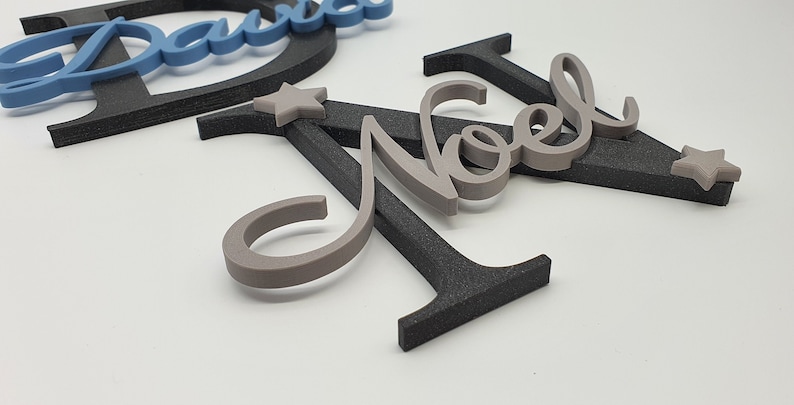 May include: Gray wooden letters spelling "Noel" with two small gray stars. The letters are layered on top of a black wooden letter "A".