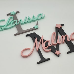 May include: Two wooden names, Larissa and Malina, are painted in cursive script. The letters are painted in pink, teal, and black. Each name has a small star painted on it.