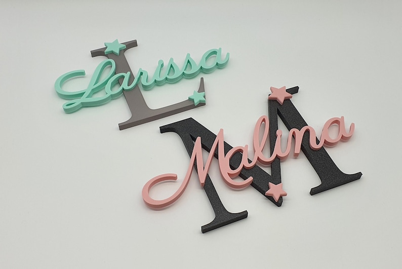 May include: Two wooden names, Larissa and Malina, are painted in cursive script. The letters are painted in pink, teal, and black. Each name has a small star painted on it.