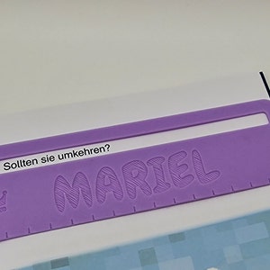 Pode incluir: Marca-p&aacute;ginas de pl&aacute;stico roxo com o nome "MARIEL" em relevo em uma fonte de bolhas. O marca-p&aacute;ginas tamb&eacute;m tem um pequeno &iacute;cone de coroa e o texto "Sollten sie umkehren?" impresso nele.
