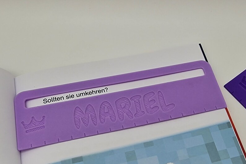 Pode incluir: Marca-p&aacute;ginas de pl&aacute;stico roxo com o nome "MARIEL" em relevo em uma fonte de bolhas. O marca-p&aacute;ginas tamb&eacute;m tem um pequeno &iacute;cone de coroa e o texto "Sollten sie umkehren?" impresso nele.
