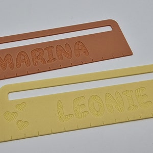Pode incluir: Duas r&eacute;guas personalizadas, uma laranja e outra amarela, com os nomes "Marina" e "Leonie" gravados nelas. Cada r&eacute;gua tem tr&ecirc;s pequenos cora&ccedil;&otilde;es na parte inferior.