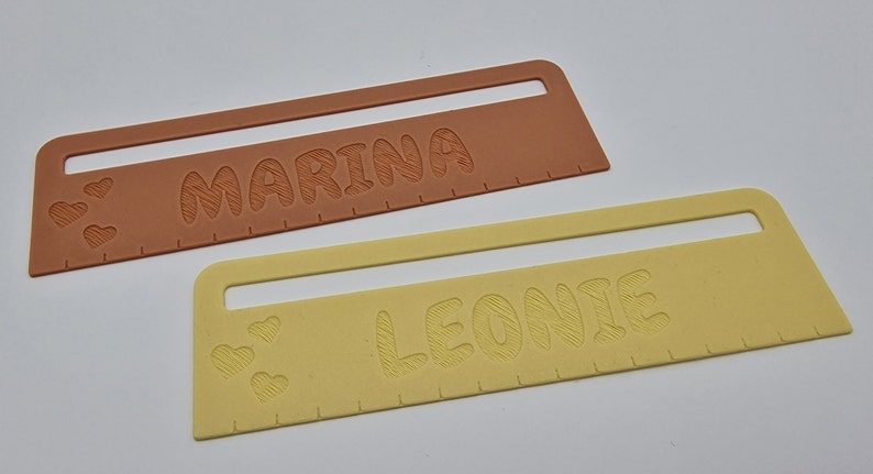 Pode incluir: Duas r&eacute;guas personalizadas, uma laranja e outra amarela, com os nomes "Marina" e "Leonie" gravados nelas. Cada r&eacute;gua tem tr&ecirc;s pequenos cora&ccedil;&otilde;es na parte inferior.