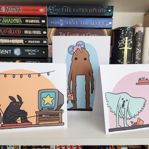 May include: Three illustrated greeting cards with different designs. The first card features two cartoon monsters playing a video game. The second card features a brown monster holding a bag. The third card features a blue ghost with a pink background.