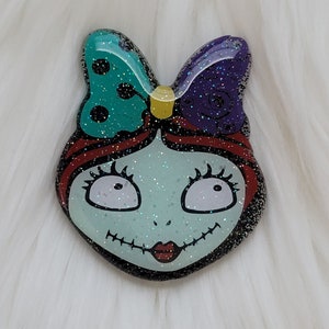 May include: A resin pin featuring a cartoon character with green, purple, and yellow bow, red hair, and a stitched smile. The character has large eyes and a light blue skin tone.