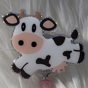 May include: A white cow with black spots, glitter, and pink and white beads on a silver retractable badge reel.