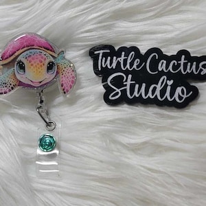 May include: A colorful, iridescent, resin turtle with a teal glitter accent is attached to a retractable badge reel. The reel has a teal glitter accent. The background is a white, fluffy surface. The text "Turtle Cactus Studio" is visible in the background.