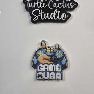 May include: A black and white Turtle Cactus Studio logo above a blue and silver Game Over controller design. The controller has a hand holding it, with the words "GAME OVER" below. The background is white.