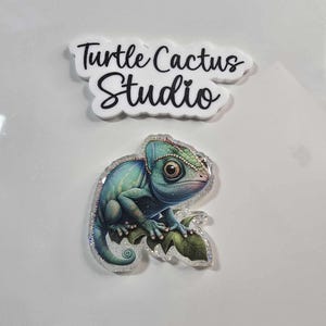 May include: A blue and green chameleon with glitter accents, perched on a leaf. The chameleon is a resin charm with a white background. The text "Turtle Cactus Studio" is printed on a white background.