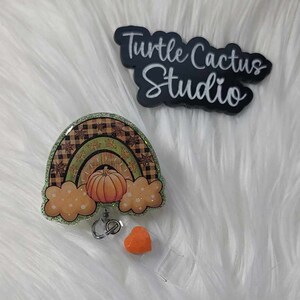 May include: A green, orange, and brown fall-themed rainbow with a pumpkin in the center. The rainbow is attached to a clear badge reel with an orange heart charm. The badge reel is on a white fuzzy surface. The text "Turtle Cactus Studio" is visible in the background.