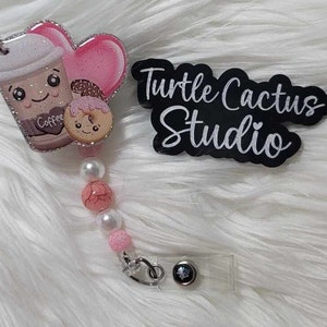 May include: A pink and brown coffee cup with a donut charm and a pink heart charm. The charm is attached to a white and pink beaded retractable badge reel with a silver clip. The badge reel is on a white fuzzy surface. The text "Turtle Cactus Studio" is on a black sign.
