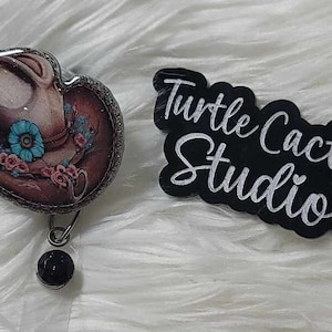 May include: A brown and white cowboy hat with blue flowers and a black bead on a silver chain. The hat is on a white fluffy surface. The text "Turtle Cactus Studio" is on a black background.