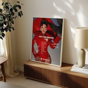 Może przedstawiać: Oprawiony plakat z napisem "AM I THE DRAMA?" z Cardi B w czerwonym stroju. Plakat zawiera listę utworów i szczegóły wydania. Grafika jest wyświetlana na drewnianej szafce, z lampą i rośliną w tle.