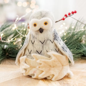 May include: A white felt owl with black eyes and a gray felt leaf in its talons. The owl is sitting on a wooden surface with a green pine branch and red berries in the background.