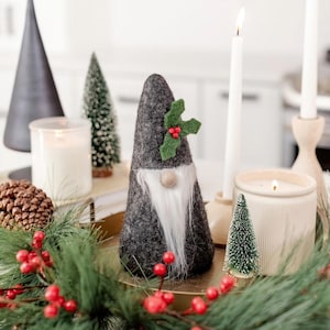 Op de afbeelding: Een grijze vilten kabouter met een witte baard, houten neus en een groene hulst accent. De kabouter maakt deel uit van een kerstdisplay met kaarsen, dennennaalden en kleine kerstbomen.