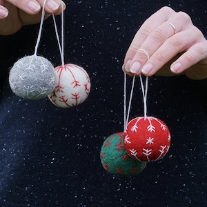 May include: Four felt Christmas ornaments with white string hangers. The ornaments are decorated with white snowflake designs. One ornament is gray, one is white, one is red, and one is green.