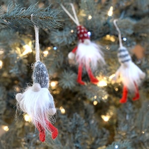 May include: Three gray and white knitted gnome ornaments with red hats and legs hanging on a Christmas tree.