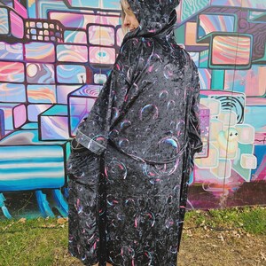 May include: A black hooded robe featuring a repeating pattern of iridescent bubbles in shades of pink, blue, and purple. The robe has long sleeves, a tie waist belt, and two front pockets, crafted from a soft, textured fabric with a slight sheen.
