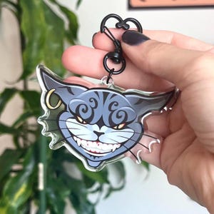 Op de afbeelding: Een heldere acryl sleutelhanger met een grijnzende cartoon kattenkop met grijze en blauwe strepen. De kat heeft gele ogen, een gouden oorbel en een zwarte sluiting. De sleutelhanger wordt door een persoon vastgehouden.