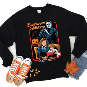May include: Black sweatshirt with a graphic print of a horror movie scene. The graphic features a masked figure, a woman, and a child. The text on the graphic reads "Halloween Safety A Sitter's Guide".