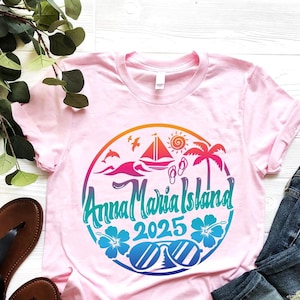 May include: Pink t-shirt with a colorful graphic design of a beach scene with a sailboat, palm trees, sunglasses, and the text "Anna Maria Island 2025".