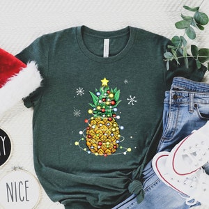 May include: Green t-shirt featuring a pineapple decorated as a Christmas tree with lights and a star. The image includes a Santa hat, the words "NAUGHTY" and "NICE" on small round signs, blue jeans, and white sneakers.