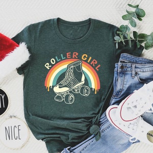 Peut inclure: Un t-shirt vert avec le texte "Roller Girl" dans une arche arc-en-ciel avec un graphique de patins &agrave; roulettes. Le t-shirt est pos&eacute; sur une surface blanche avec un chapeau de P&egrave;re No&euml;l, deux pancartes rondes qui disent "Naughty" et "Nice", un jean bleu et des baskets blanches.