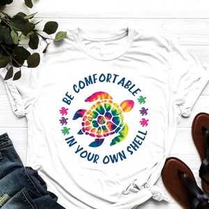 May include: White t-shirt with a tie-dye turtle graphic and the text "BE COMFORTABLE IN YOUR OWN SHELL" in blue. The shirt is paired with blue jeans and black sandals. A sprig of green leaves is in the background.