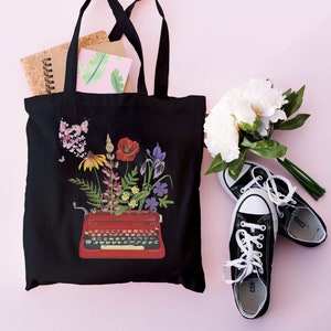 May include: Black tote bag with a red typewriter with flowers coming out of it. The bag is on a pink surface with a bouquet of white flowers and black sneakers.