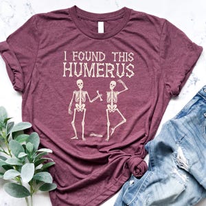 May include: Burgundy t-shirt with the text "I FOUND THIS HUMERUS" above a graphic of two skeletons. One skeleton is holding a bone, and the other is posing. Light blue denim jeans are next to the shirt.