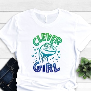 May include: White t-shirt with a green, blue, and purple gradient design. The design features a cartoon dinosaur with the text "Clever Girl" above it.
