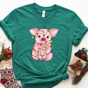May include: A teal green t-shirt with a pink pig wearing Christmas lights. The pig is sitting down with its legs crossed and has a happy expression on its face.