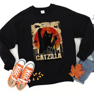 May include: Black sweatshirt with a graphic of a black cat with the text "CATZILLA" and Japanese characters. The cat is in a fighting pose with its claws out. The background is a red and orange sunset with a cityscape.