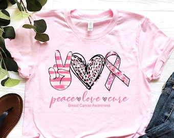 May include: Pink t-shirt with a peace sign, heart, and pink ribbon with the text "peace love cure" and "Breast Cancer Awareness".