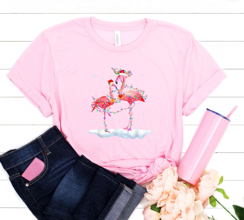 May include: Pink t-shirt with a Christmas-themed flamingo design. The flamingos are wearing Santa hats and have Christmas lights wrapped around them. The design is on a pink t-shirt with a white background.