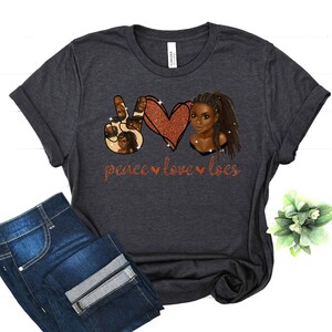 May include: A dark gray t-shirt with a peace sign, heart, and a woman with locs. The design is in glitter and says "peace love locs".