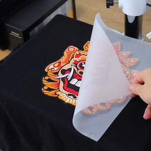 May include: A black t-shirt with a colorful graphic design being revealed. The design features a detailed, ornate illustration with red, orange, yellow, and white accents. A hand is lifting a translucent sheet to reveal the design.